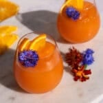 A side shot of Two stemless wine glasses with Aperol Spritz slushies topped with orange wedges and edible purple flowers.