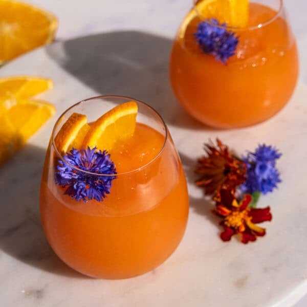 A side shot of Two stemless wine glasses with Aperol Spritz slushies topped with orange wedges and edible purple flowers.