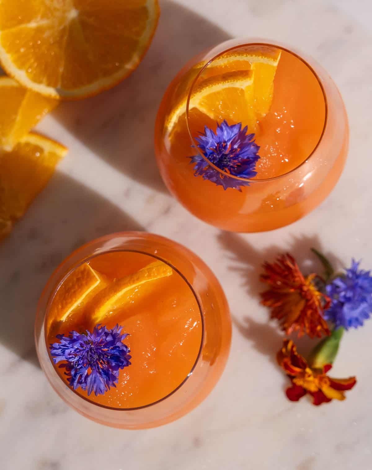 Two Aperol Spritz Slushies in wine glasses, garnished with edible flowers and orange slices. 