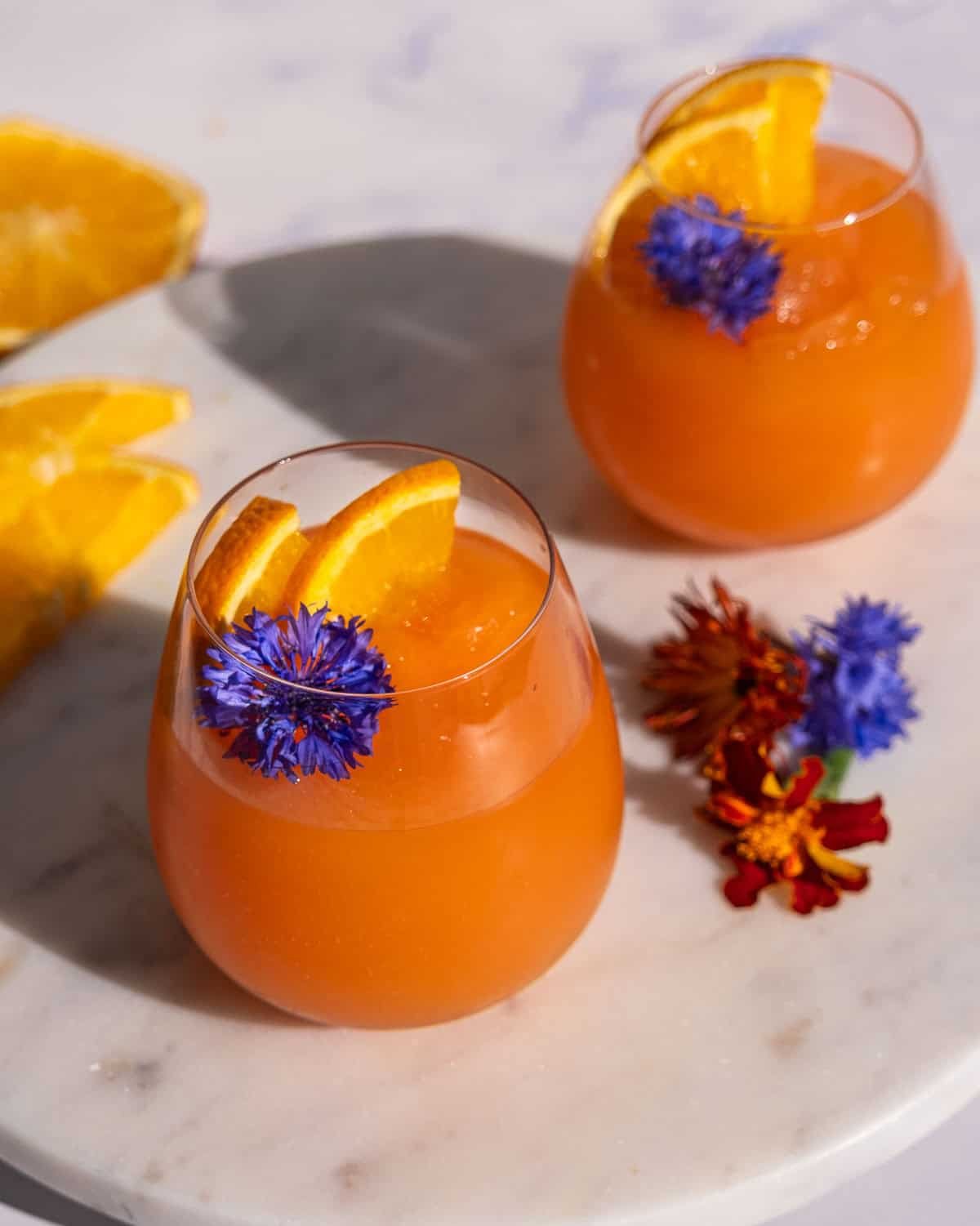 Two stemless wine glasses filled with Aperol Spritz Slushies garnished with flowers and orange peel. 