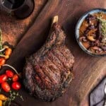 A top down image of a grilled cowboy ribeye steak on a wooden cutting board and a side of mushroom sauce.