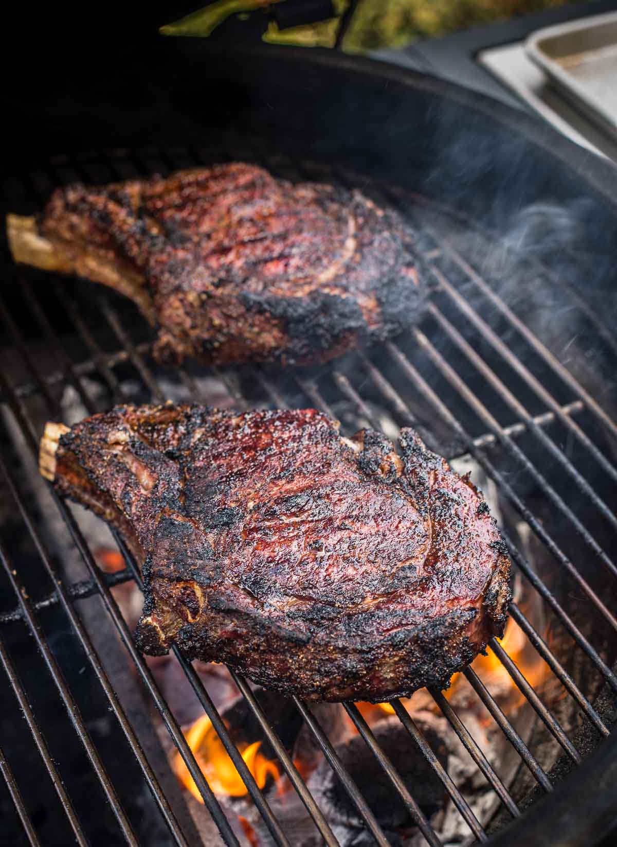 Two cowboy ribeye steaks cooking on a grill over an open flame.