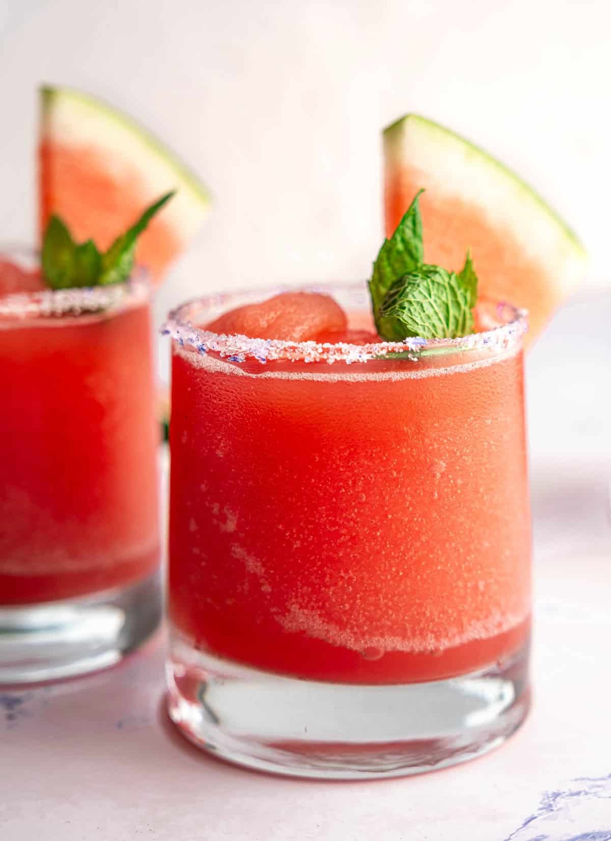 A glass filled with a Watermelon Frosé Slushy garnished with a slice of watermelon and sprig of mint.