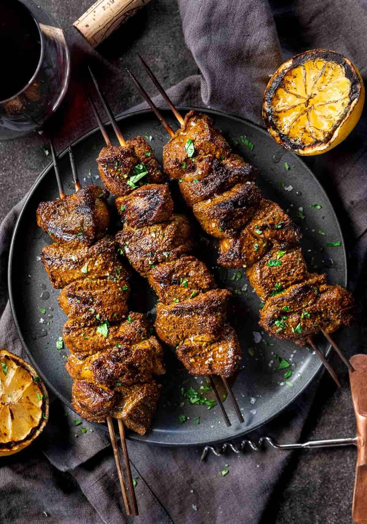Grilled Moroccan Lamb Skewers on a serving platter with a side of grilled lemon and a glass of wine.