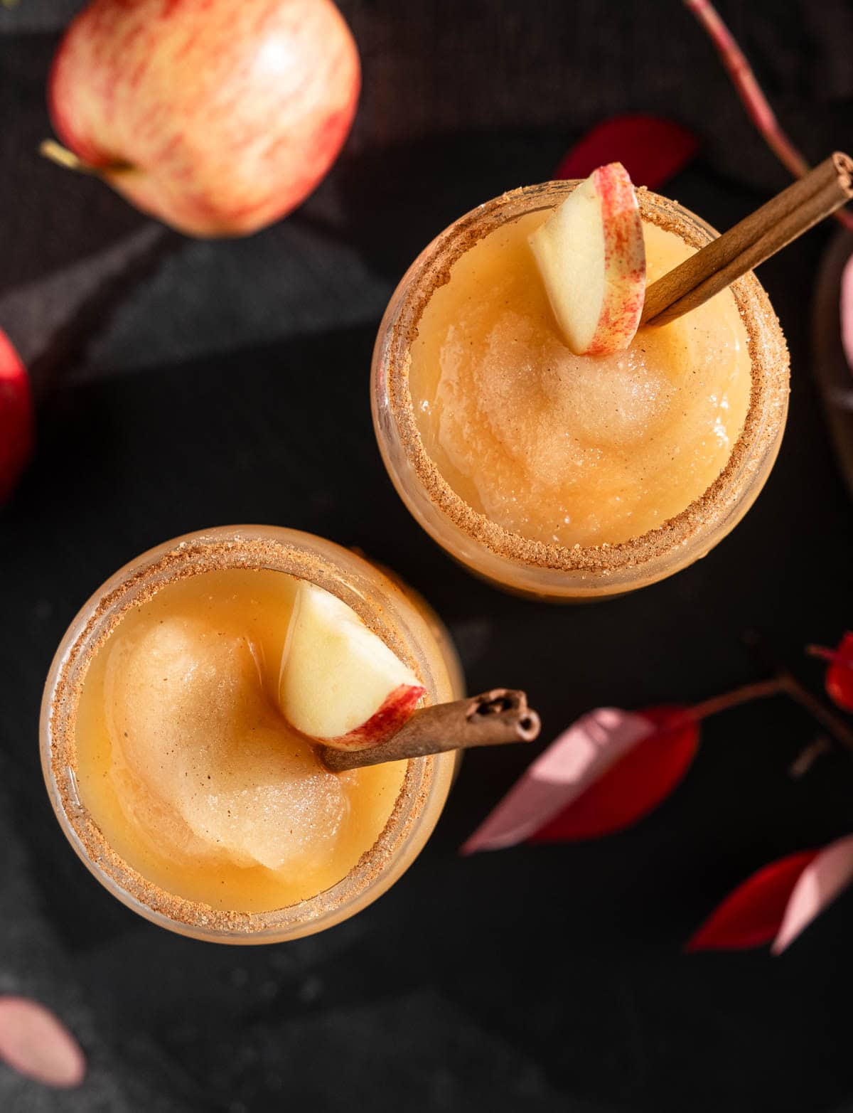 A top-down view of two spiced apple cider slushies, rimmed with cinnamon sugar and garnished with apple slices and a cinnamon stick.