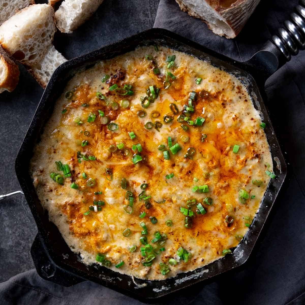 Crab dip with Dungeness crab and smoked salmon in a large cast iron pan surrounded by fresh slices of bread.