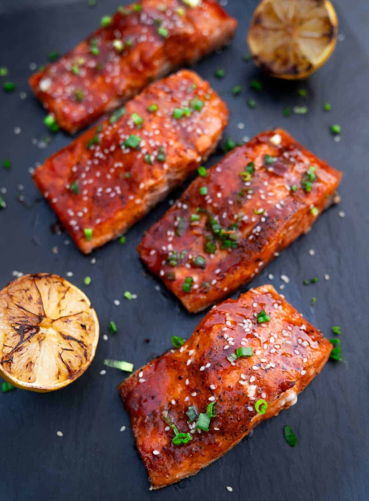 Four filets of grilled salmon topped with Gochujang sauce on a black platter with a slice of grilled lemon. 