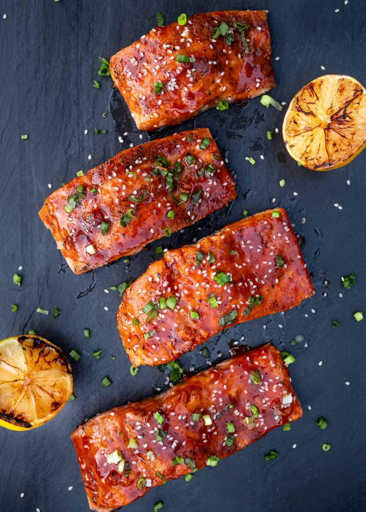 Four grilled salmon filets coated with a savory Gochujang Sauce glaze on a serving platter with a slice of grilled lemon. 