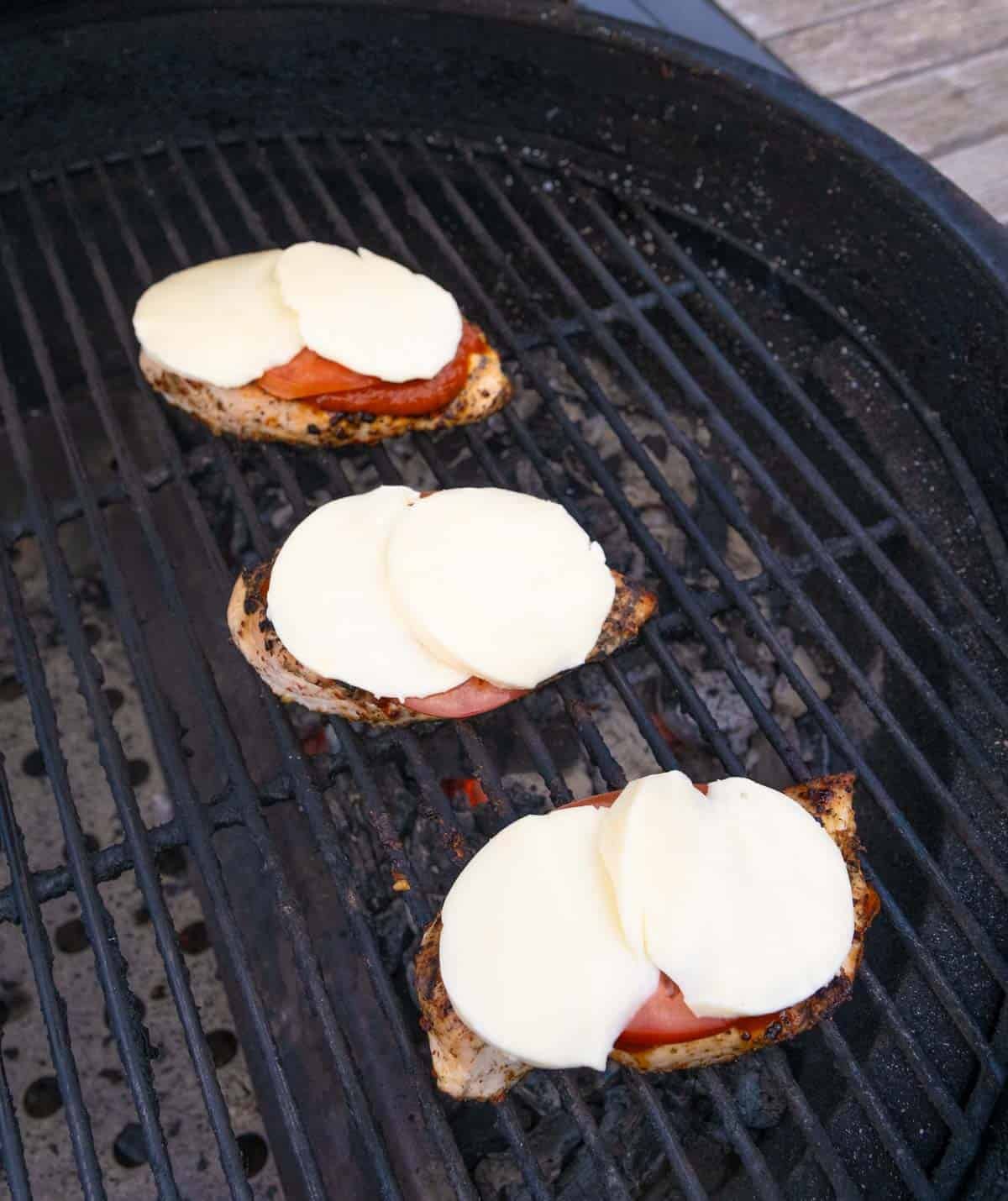 Three chicken breasts cooking on the grill topped with mozzarella cheese.