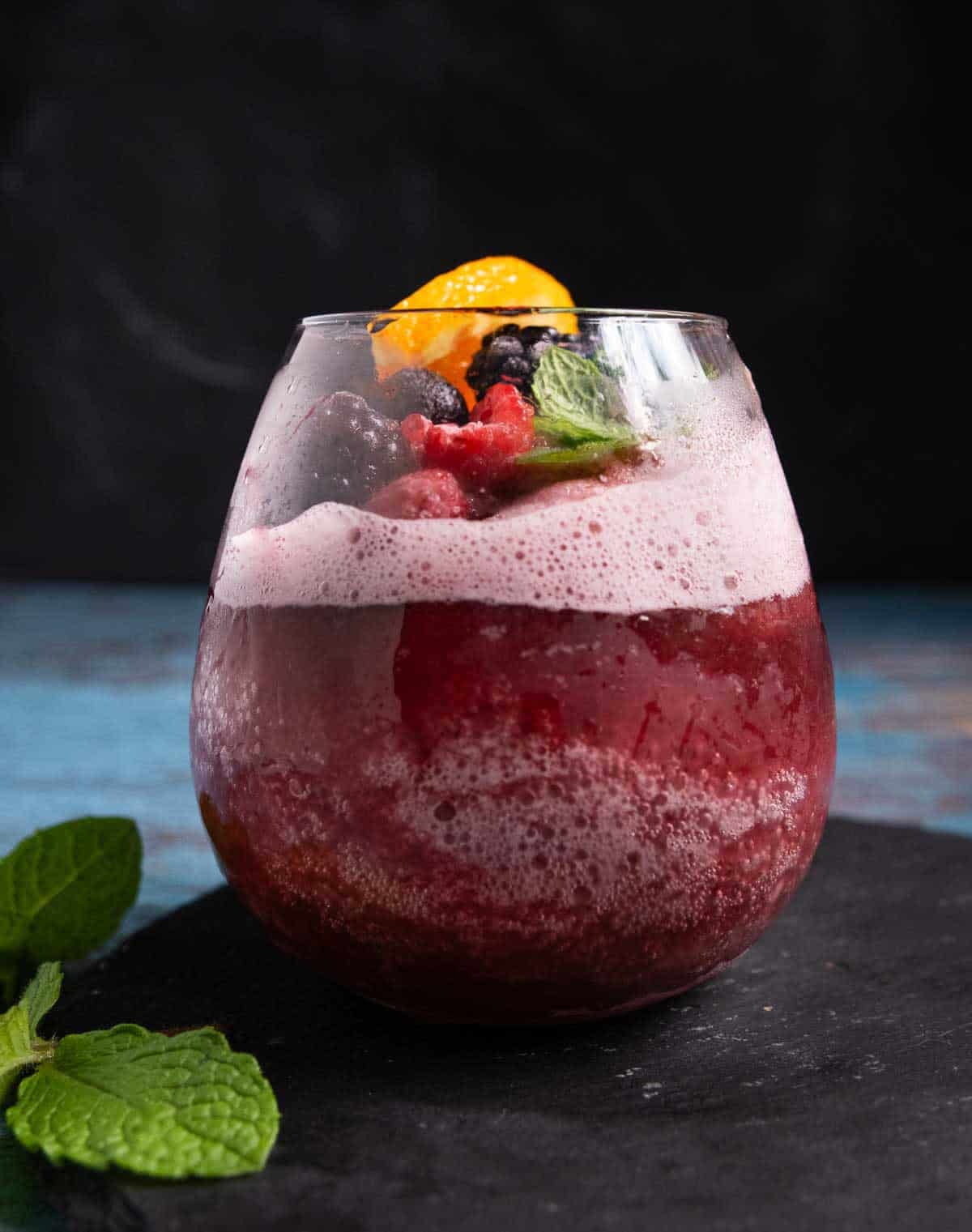 A frozen red wine sangria slushie in a stemless wine glass topped with frozen berries and a sprig of mint.