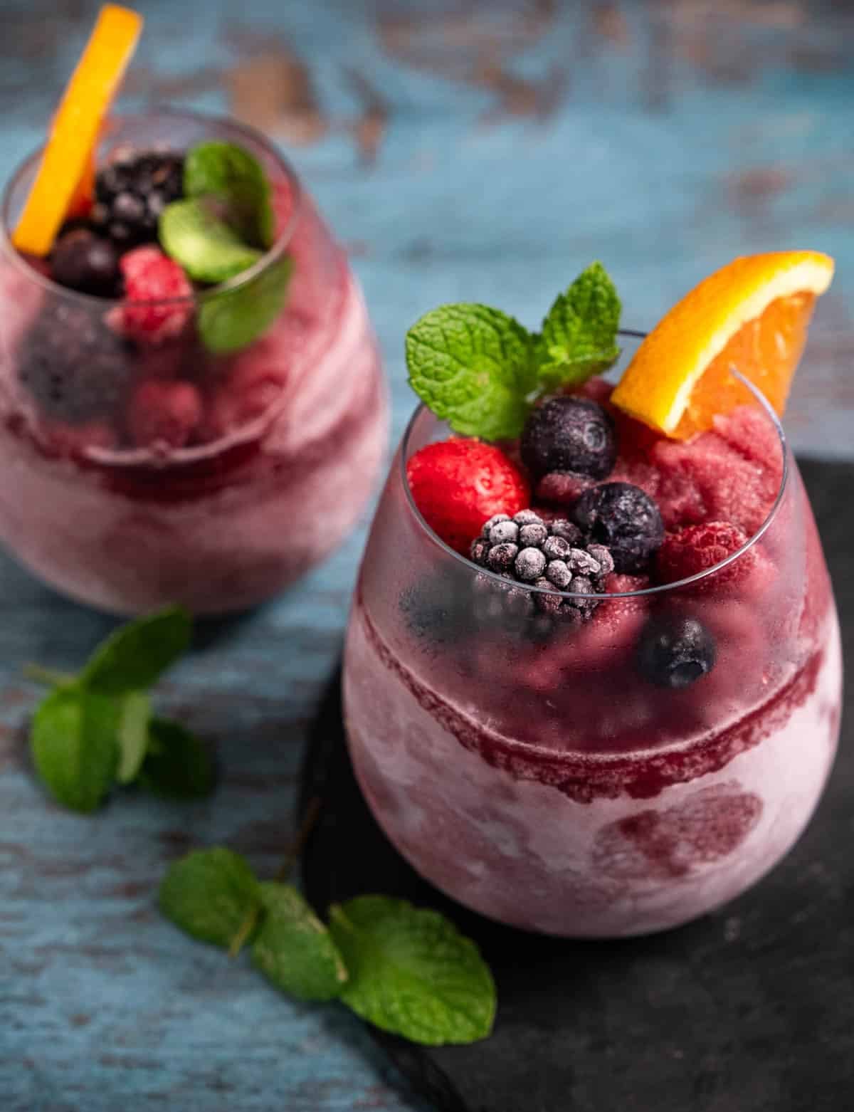 Two stemless glasses filled with red wine sangria slushies made in the Ninja Slushi Machine, and garnished with frozen berries, mint and orange slices.