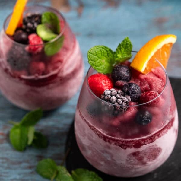 Two red wine slushies made in a Ninja Slushi Machine, served in stemless wine glasses, and garnished with frozen berries and an orange slice.