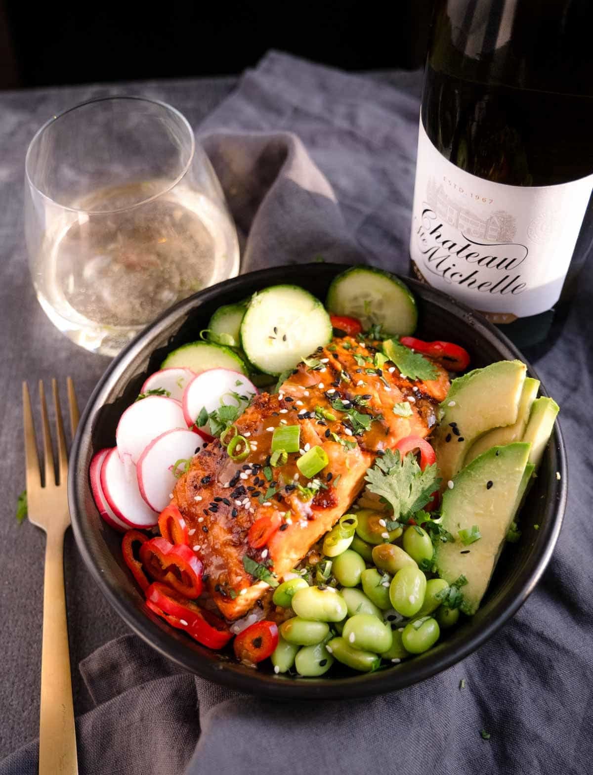 Grilled miso salmon rice bowl with avocado, edamame, cucumber, and radishes with a bottle of Riesling next to the salmon bowls.
