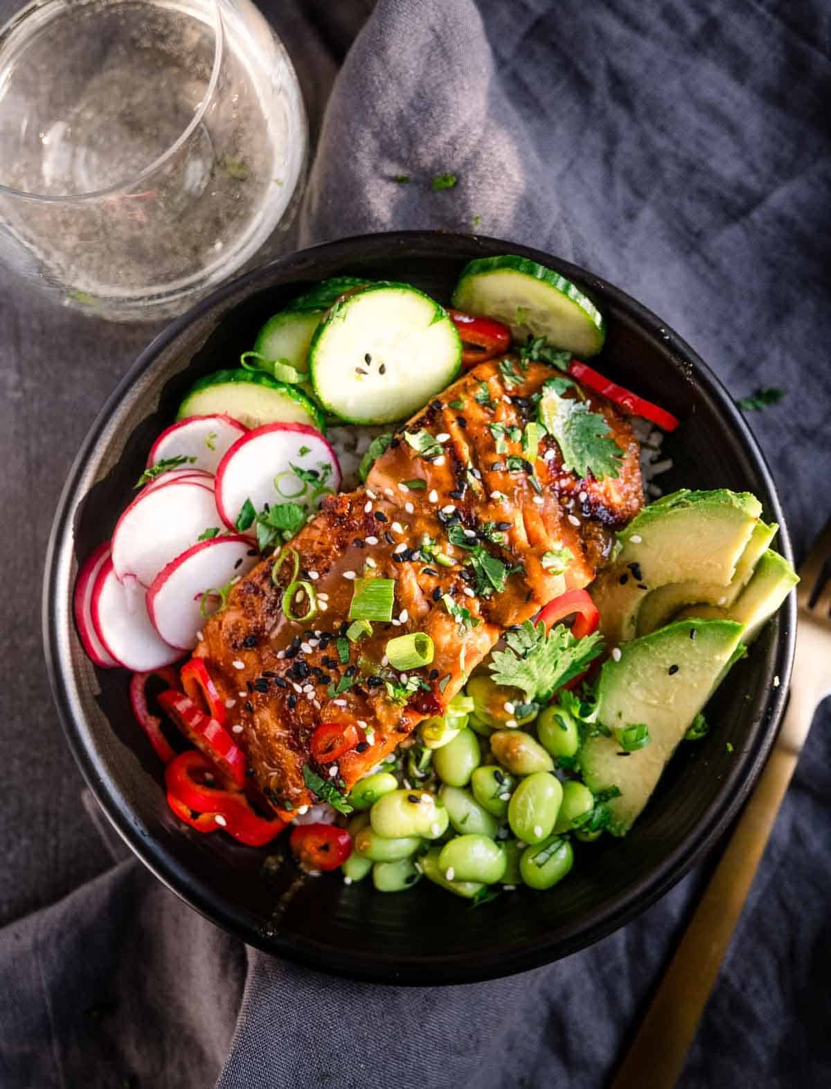 A bowl of grilled miso-glazed salmon served over rice, alongside cucumber slices, radishes, edamame, and avocado slices.